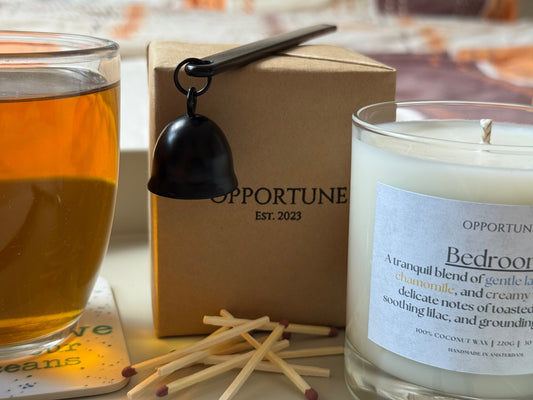 A tray placed on a bed. On tray, you have a glass of tea, a boxed candle, an unboxed candle, a wick snuffer and some scattered matches.
